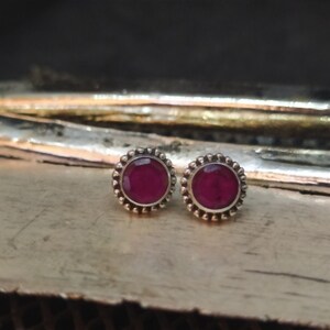 May include: A pair of silver stud earrings with a round, dark red gemstone in the center of each earring. The earrings have a decorative border of small silver beads.