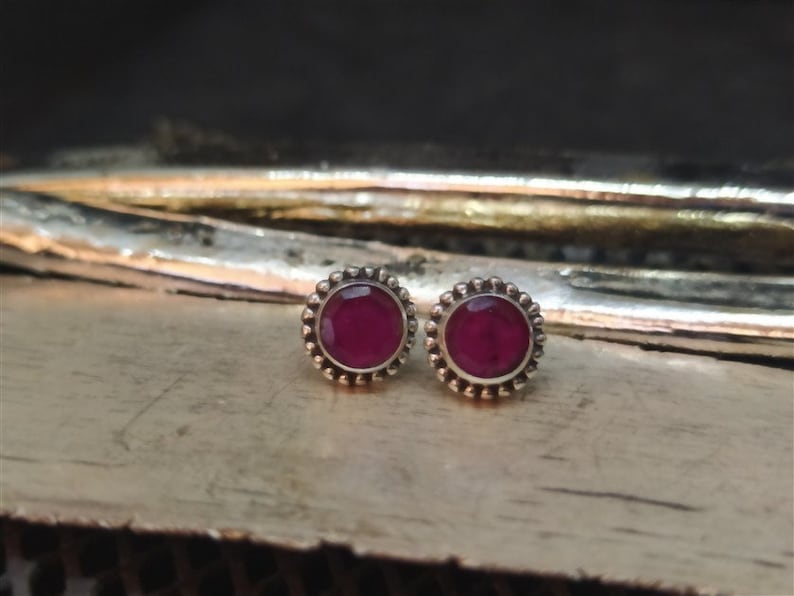 May include: A pair of silver stud earrings with a round, dark red gemstone in the center of each earring. The earrings have a decorative border of small silver beads.