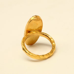 Może przedstawiać: Złoty pierścionek z dużym, owalnym kamieniem szlachetnym. Obrączka pierścionka jest fakturowana i ma młotkowane wykończenie.