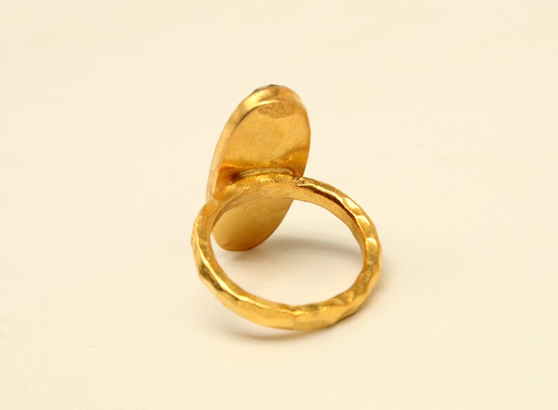 Może przedstawiać: Złoty pierścionek z dużym, owalnym kamieniem szlachetnym. Obrączka pierścionka jest fakturowana i ma młotkowane wykończenie.