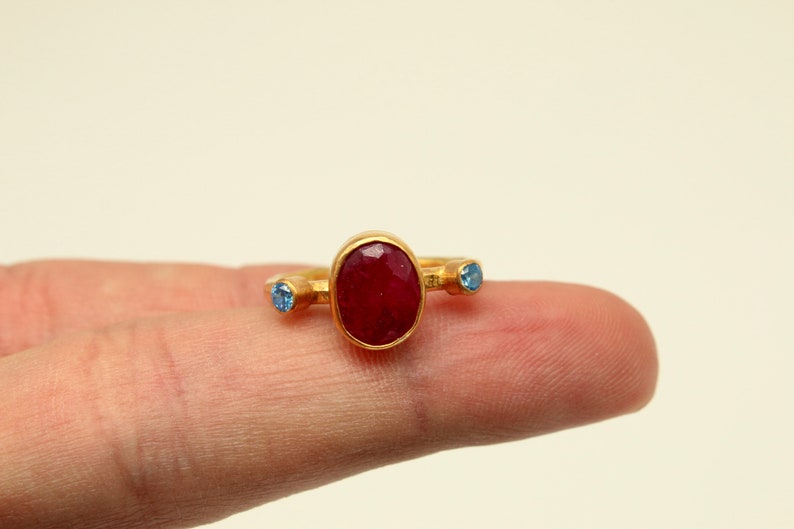 Peut inclure: Une bague en or avec une grande pierre rouge ovale et deux petites pierres bleues de chaque c&ocirc;t&eacute;.