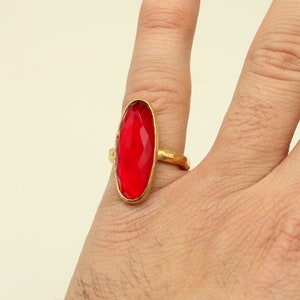 May include: A gold ring with a large, oval-shaped, red gemstone. The ring is worn on a person's finger.