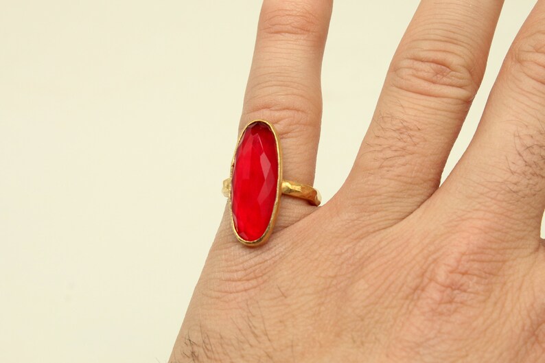 May include: A gold ring with a large, oval-shaped, red gemstone. The ring is worn on a person's finger.