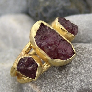 May include: A gold ring with three raw ruby stones set in the band. The stones are a deep red color and have a rough, natural texture.