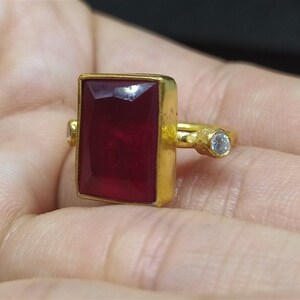 Op de afbeelding: Een gouden ring met een grote, rechthoekige, donkerrode edelsteen en een kleine, ronde, heldere edelsteen.