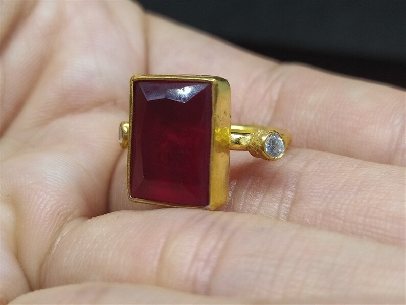 Op de afbeelding: Een gouden ring met een grote, rechthoekige, donkerrode edelsteen en een kleine, ronde, heldere edelsteen.