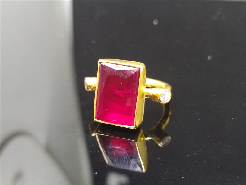 Op de afbeelding: Een gouden ring met een grote, rechthoekige, rode edelsteen en een kleinere, ronde, heldere edelsteen aan de zijkant.