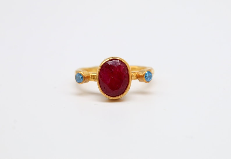 Peut inclure: Une bague en or avec une grande pierre de rubis ovale et deux petites pierres bleues de chaque c&ocirc;t&eacute;.