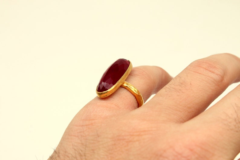 Może przedstawiać: Złoty pierścionek z dużym, owalnym, ciemnoczerwonym kamieniem szlachetnym.