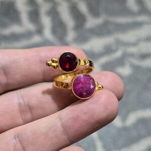 May include: A gold-toned bypass ring featuring two round, faceted gemstones. The top stone is a deep, dark red, while the bottom stone is a vibrant, opaque magenta. The metal band has a textured, hammered finish with small decorative bead accents.