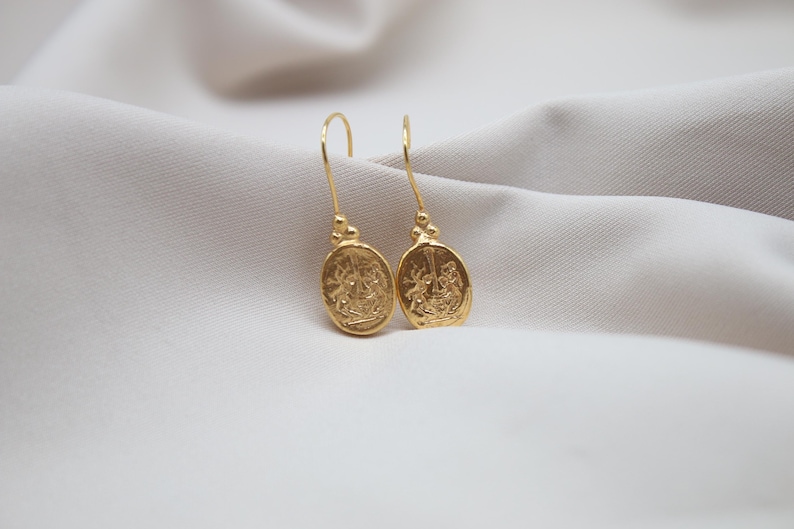 May include: A pair of gold-toned dangle earrings with oval-shaped charms featuring intricate embossed designs.