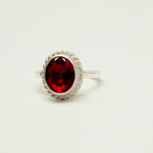 Op de afbeelding: Een zilveren ring met een rode ovale edelsteen gezet in een touwachtige rand.