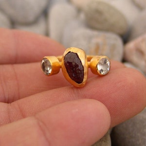 May include: Gold ring featuring a large, dark red gemstone and two smaller, clear gemstones.