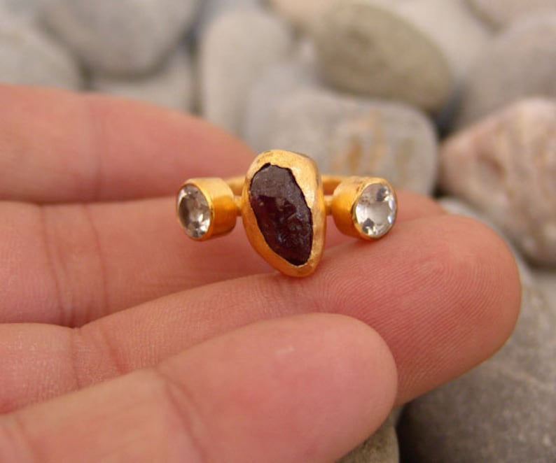 May include: Gold ring featuring a large, dark red gemstone and two smaller, clear gemstones.