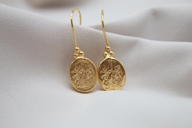 May include: A pair of gold dangle earrings with a detailed oval design featuring a raised image of two figures.
