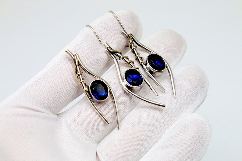 May include: A pair of silver earrings with blue gemstones and gold accents. The earrings are shaped like a teardrop and have a delicate, intricate design.