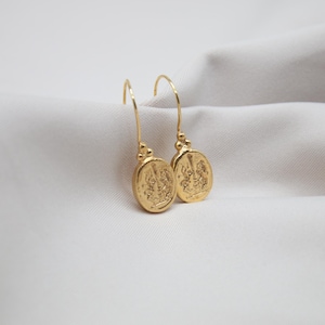 May include: A pair of gold dangle earrings with oval-shaped charms featuring intricate designs. The charms are suspended from delicate gold hooks.