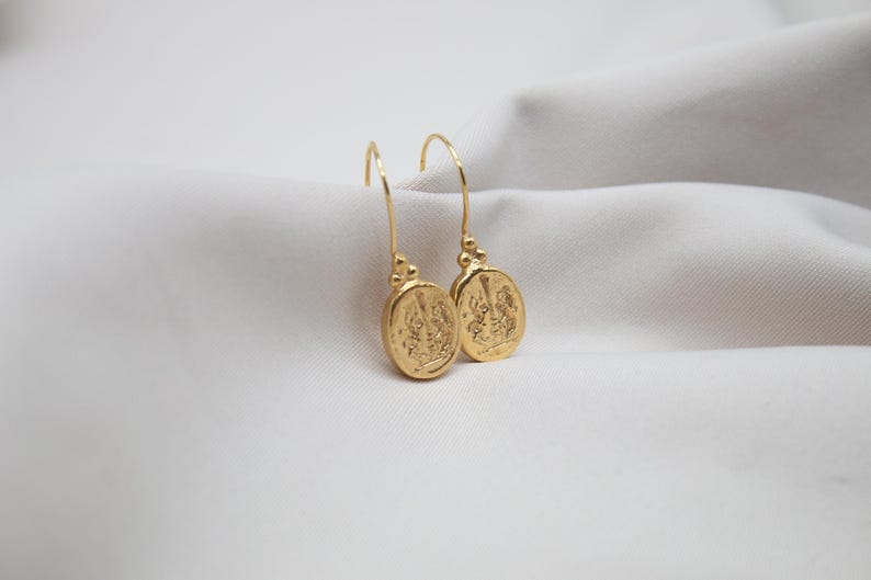 May include: A pair of gold dangle earrings with oval-shaped charms featuring intricate designs. The charms are suspended from delicate gold hooks.