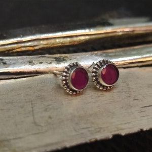 May include: A pair of silver stud earrings with small, round, dark pink gemstones set in a beaded bezel.