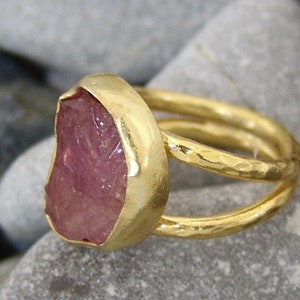 Pode incluir: Um anel de ouro com uma gema rosa engastada em um engaste de bezel. O anel tem um acabamento martelado e uma banda dupla.
