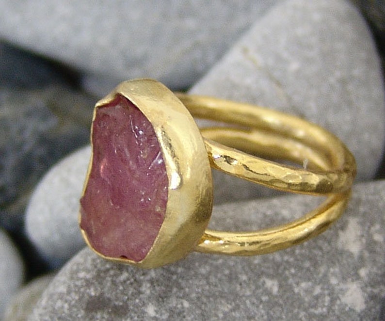 Pode incluir: Um anel de ouro com uma gema rosa engastada em um engaste de bezel. O anel tem um acabamento martelado e uma banda dupla.