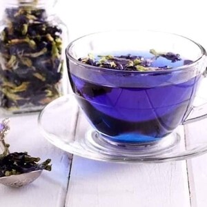 May include: A glass cup filled with blue butterfly pea flower tea. The tea is a vibrant blue color and there are some flower petals floating in the tea. The cup is sitting on a white saucer.