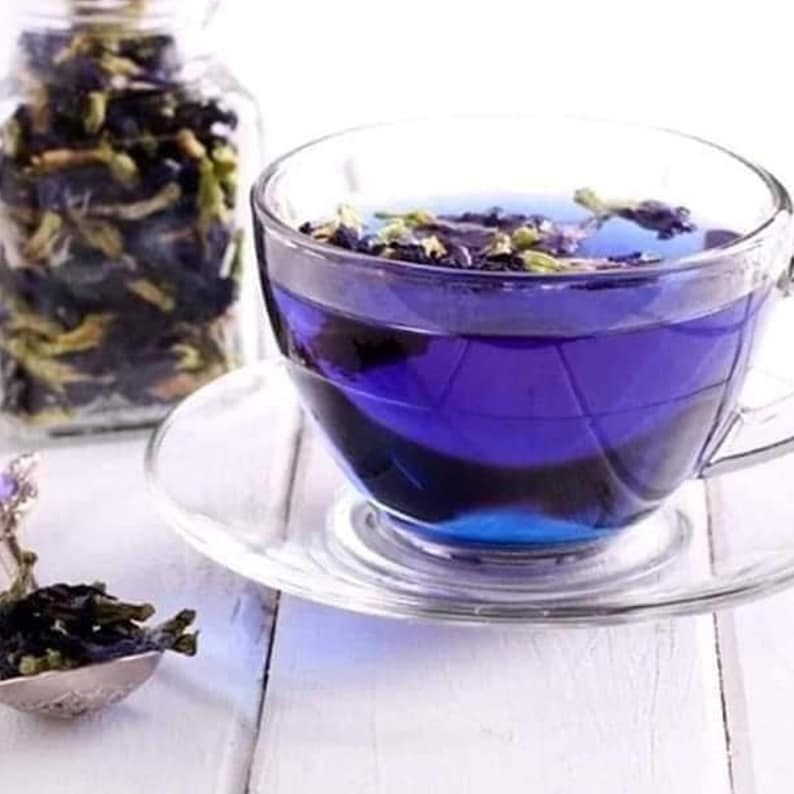 May include: A glass cup filled with blue butterfly pea flower tea. The tea is a vibrant blue color and there are some flower petals floating in the tea. The cup is sitting on a white saucer.
