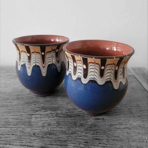 May include: Two blue ceramic mugs with brown and white painted designs. The mugs have a rounded shape and a wide rim.