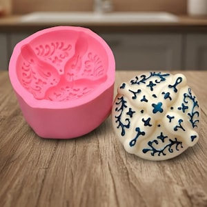 Puede incluir: Un molde de silicona rosa redondo con un intrincado patrón en relieve de un pájaro y flores, junto a un objeto circular blanco terminado con un diseño de cruz pintado en azul y detalles decorativos de filigrana.