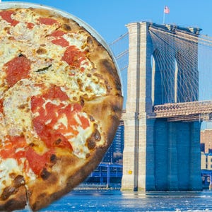 Pode incluir: Uma pizza grande com pepperoni e queijo numa bandeja de metal. A pizza está em frente à Ponte do Brooklyn em Nova York, com um céu azul e a bandeira americana visível.