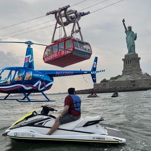 Pode incluir: Uma pessoa numa moto aquática Yamaha branca e amarela na água, com um helicóptero azul e um teleférico vermelho de Roosevelt Island ao fundo. A Estátua da Liberdade é visível ao longe.