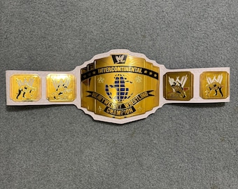 Puede incluir: Un cinturón de campeonato de lucha libre blanco y dorado con las palabras "Intercontinental Heavyweight Wrestling Champion" y un diseño de globo terráqueo. El cinturón tiene placas laterales con figuras de lucha libre.