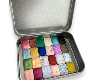 May include: A silver metal tin containing 24 watercolor paint pans in various colors. The colors include shades of brown, gold, blue, pink, and green. The tin is open, revealing the rectangular paint pans.