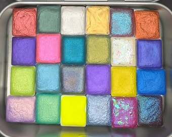Könnte beinhalten: Eine Metalldose gefüllt mit 20 kleinen Quadraten Aquarellfarbe. Die Farbe ist in verschiedenen Farben, darunter Rot, Grün, Blau, Gelb, Lila und Pink. Die Dose ist geöffnet und die Farbe ist sichtbar.
