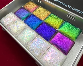 Könnte beinhalten: Eine silberne Metalldose mit einem durchsichtigen Plastikdeckel, die 12 kleine Quadrate mit schimmernder Aquarellfarbe enthält. Die Dose trägt den Aufdruck "Colourshift Countdown to Christmas". Die Farben der Farbe umfassen Gold, Grün, Blau, Lila und Weiß.