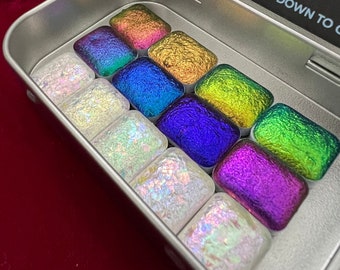 Puede incluir: Una lata de metal plateada con una tapa negra que contiene 12 pequeños cuadrados de pintura iridiscente. La tapa tiene las palabras "Colourshift Countdown to Christmas" impresas. Los cuadrados de pintura están dispuestos en una cuadrícula y son de diferentes colores.