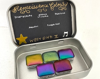 May include: A silver metal tin with a black lid. The lid has the words "Renaissance Colours" written in gold cursive. The tin contains six small squares of iridescent paint. The paint squares are labeled with the names "Shaftesbury", "Apollo Victoria", "Peacock", "Adelphi", "Cambridge", and "West End II".