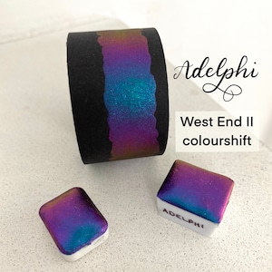 May include: A close-up of a black and iridescent watercolor pan with the text "Adelphi West End II colourshift" on a white surface. The pan is a small square shape and has a metallic finish.
