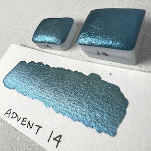 Puede incluir: Dos pastillas de pintura de acuarela, una m&aacute;s grande que la otra, ambas etiquetadas con el n&uacute;mero "14". La pastilla m&aacute;s grande es de color azul verdoso con un brillo met&aacute;lico. La pastilla m&aacute;s peque&ntilde;a es del mismo color. Se muestra una muestra de la pintura de acuarela azul verdosa sobre una superficie blanca con el texto "Advent 14" escrito debajo.