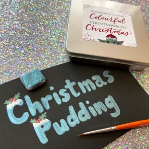 Puede incluir: Una superficie negra con las palabras "Christmas Pudding" escritas en pintura azul. Hay dos peque&ntilde;as ilustraciones de pud&iacute;n navide&ntilde;o azul y blanco con ramitas de acebo. Tambi&eacute;n se puede ver un peque&ntilde;o bote de pintura y un pincel. El texto "@justwriteawayrenee" est&aacute; escrito en blanco en la superficie.
