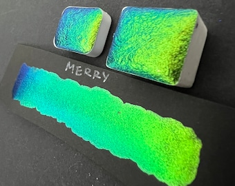 May include: Two small, square, metallic paint pans with a green and blue iridescent shimmer. The pans are labeled "Merry" in white lettering.