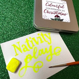 May include: A white card with the words "Nativity Play" written in yellow paint. A small yellow paint pan and a paintbrush are on the card. The card is on a green sparkly surface.