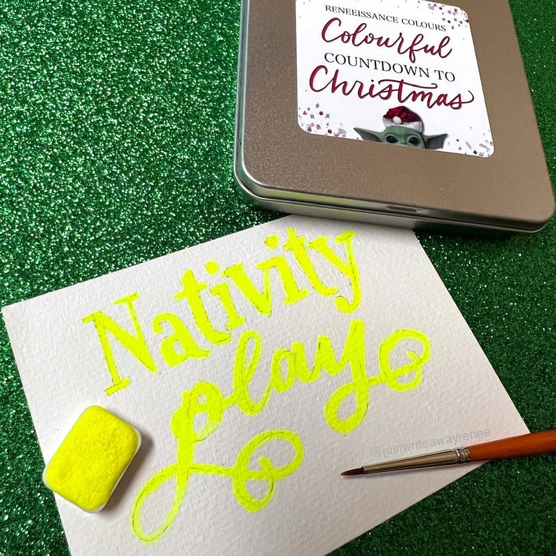 May include: A white card with the words "Nativity Play" written in yellow paint. A small yellow paint pan and a paintbrush are on the card. The card is on a green sparkly surface.