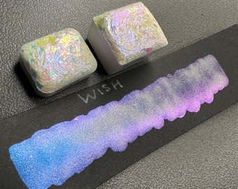 May include: Two small square containers of iridescent white paint with a shimmery finish. The paint is labeled "WISH" and is shown swiped on a black surface.