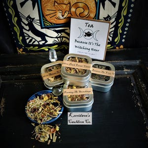 May include: An assortment of herbal teas in small metal tins and a blue bowl, with a sign that reads "Tea Because It's The Witching Hour." The tins are labeled with names like "Idren Dark Forest Coven" and "Evening At Stonehenge."