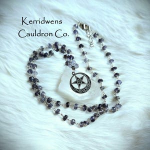 May include: A necklace featuring a pendant with a pentagram inside a crescent moon design. The necklace is made of small, dark blue and gray beads strung on a silver chain. The text "Kerridwens Cauldron Co." is visible in the image.