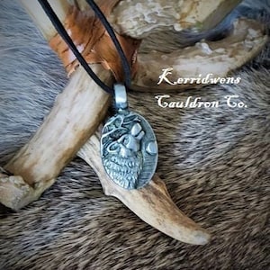 May include: A silver pendant necklace with a detailed image of a wolf's head with antlers. The pendant is oval shaped and hangs from a black cord. The pendant is on a brown fur background with the text "K.erridwens Cauldron Co."