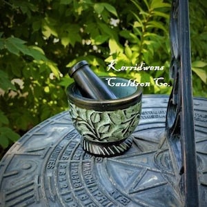 May include: A green and black mortar and pestle with a carved floral design. The mortar and pestle are sitting on a black surface with a circular design. The text "Kerridwens Cauldron Co." is visible in the background.