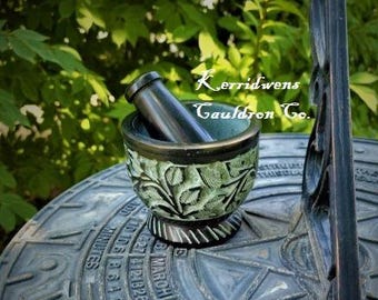 May include: A green and black mortar and pestle with a carved floral design. The mortar and pestle are sitting on a black surface with a circular design. The text "Kerridwens Cauldron Co." is visible in the background.