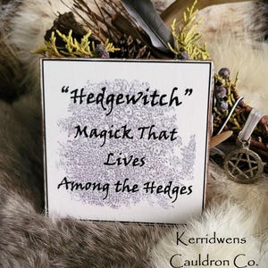 May include: White wooden sign with black text that reads "Hedgewitch" Magick That Lives Among the Hedges. The sign is on a white fur background with a feather and a pentagram charm.